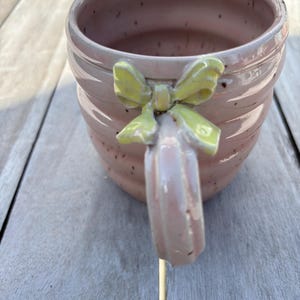 Könnte beinhalten: Eine handgefertigte rosa Keramiktasse mit einer hellgelben Schleife am Griff. Die Tasse hat ein strukturiertes, geripptes Design und ein gesprenkeltes Finish. Der Griff ist ebenfalls rosa und hat eine abgerundete Form.