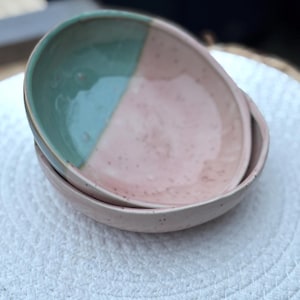 Könnte beinhalten: Zwei Keramikschalen gestapelt. Die obere Schale ist diagonal geteilt, mit einem mintgrünen Abschnitt und einem gesprenkelten rosa Abschnitt. Die untere Schale ist einfarbig gesprenkelt rosa. Die Schalen stehen auf einer weißen, gewebten Oberfläche.