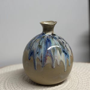 Könnte beinhalten: Eine kleine, runde Keramikvase mit beigem Sockel und einem helleren Hals. Die Vase ist mit einer blauen, weißen und grünen Glasur verziert, die an den Seiten herunterläuft und einen einzigartigen, künstlerischen Effekt erzeugt. Die Vase steht auf einer weißen Oberfläche.