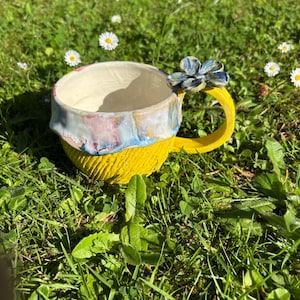 Könnte beinhalten: Ein Keramikbecher mit einer leuchtend gelben, strukturierten Basis und einem glatten, glänzenden Rand mit blauen, rosa und weißen Wirbeln. Der Henkel ist gelb und mit einer geformten, mehrblättrigen Blume in Blau-, Grau- und Weißtönen verziert.