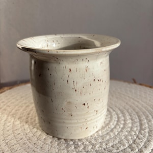 Könnte beinhalten: Ein gesprenkelter, cremefarbener Keramikbehälter mit einem breiten, leicht ausgestellten Rand. Der zylindrische Körper hat eine subtile Textur und ist mit kleinen braunen Flecken versehen. Der Behälter steht auf einer gewebten, kreisförmigen Matte.