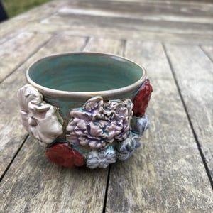 Könnte beinhalten: Eine handgefertigte Keramikschale mit türkisfarbenem Innenraum und strukturierter Außenseite, verziert mit geformten Blumen in Weiß, Rot und Lila. Die Schale steht auf einer verwitterten Holzoberfläche.