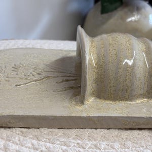 May include: A handmade, off-white ceramic soap dish with a built-in holder. The dish features a floral design and a small hole. The holder has a curved, ribbed design. The piece is textured and has a rustic aesthetic.