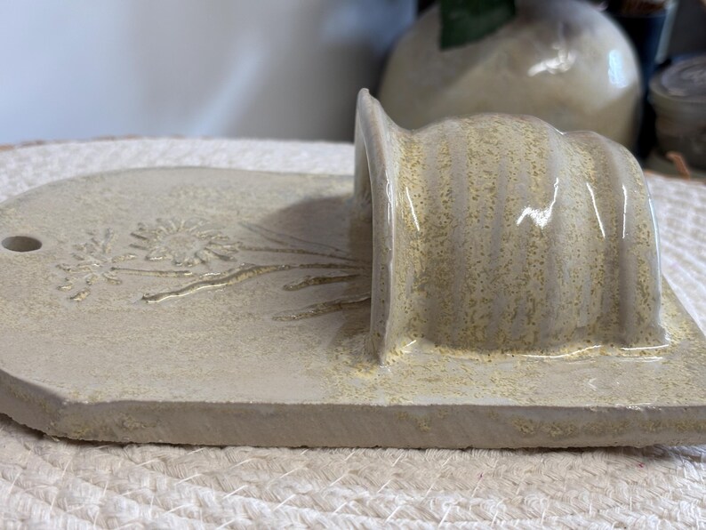 May include: A handmade, off-white ceramic soap dish with a built-in holder. The dish features a floral design and a small hole. The holder has a curved, ribbed design. The piece is textured and has a rustic aesthetic.