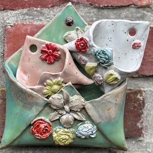 Könnte beinhalten: Eine keramische Wandtasche in Form eines Umschlags, mit einer tealgrünen Außenseite und einer rosa Innenseite. Der Umschlag ist mit bunten Keramikblumen verziert, darunter rote, gelbe und blaue Rosen sowie eine große silberne Orchidee.