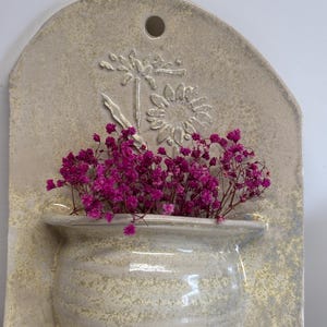 May include: A wall-mounted ceramic planter with a light beige finish. The planter features a textured, rounded pot shape filled with vibrant pink dried flowers. A raised floral design is visible on the backing.