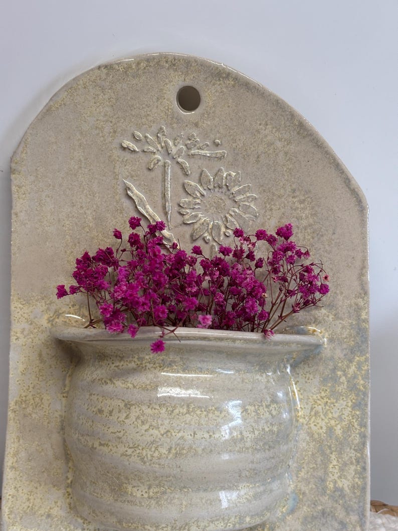 May include: A wall-mounted ceramic planter with a light beige finish. The planter features a textured, rounded pot shape filled with vibrant pink dried flowers. A raised floral design is visible on the backing.