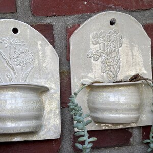 May include: Two off-white ceramic wall planters with embossed floral designs. Each planter has a small, rounded pot attached to a rectangular backboard. One features a daisy, the other a floral bouquet. The planters are mounted on a red brick wall.