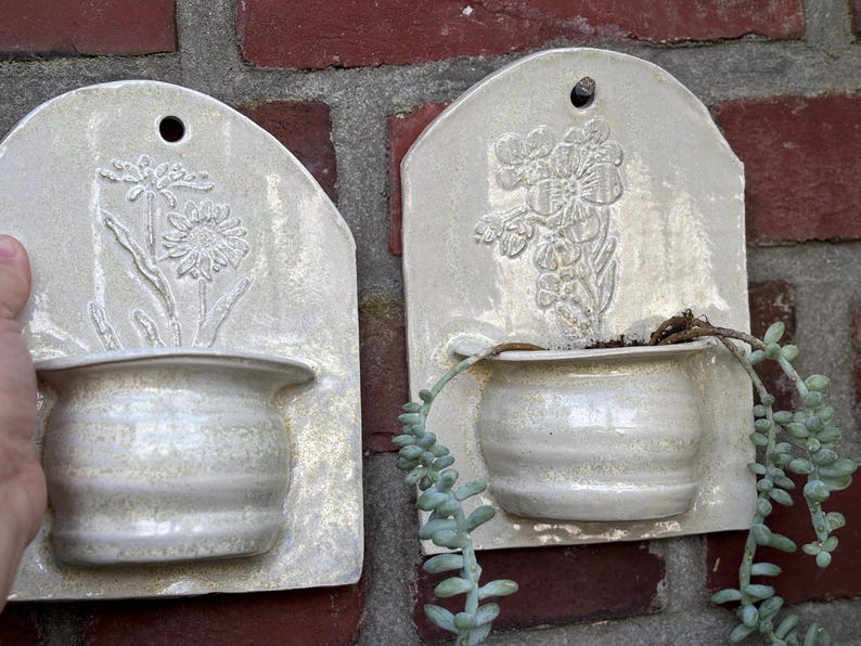 May include: Two off-white ceramic wall planters with embossed floral designs. Each planter has a small, rounded pot attached to a rectangular backboard. One features a daisy, the other a floral bouquet. The planters are mounted on a red brick wall.