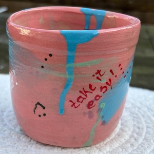 Könnte beinhalten: Eine handbemalte Keramiktasse mit rosa Grundfarbe, blauen Tropfen und den Worten "take it easy!" in Rot. Die Tasse hat ein Smiley-Gesicht und schwarze Punkte. Die Tasse ist ein einzigartiges Kunstwerk.