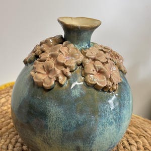 Könnte beinhalten: Eine Keramikvase mit rundem Körper und schmalem Hals. Die Vase ist in Türkis- und Blautönen glasiert, mit beigefarbenen floralen Verzierungen am oberen Rand. Der Rand ist hellbeige. Die Vase steht auf einer gewebten Oberfläche.
