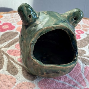 Könnte beinhalten: Ein handgefertigter keramikfroschförmiger Behälter in Grüntönen. Der Frosch hat einen weit geöffneten Mund und zwei kleine Löcher für die Augen. Der Behälter steht auf einer Oberfläche mit floralem Muster.