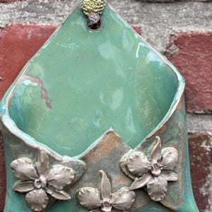Könnte beinhalten: Eine türkisfarbene Keramik-Wandtasche mit drei silberfarbenen Blumenverzierungen. Das handgefertigte Töpferstück hat eine dreieckige Oberseite mit einem kleinen, dekorativen goldfarbenen Akzent und einem Loch zum Aufhängen. Die Wandtasche ist an einer roten Backsteinmauer befestigt.