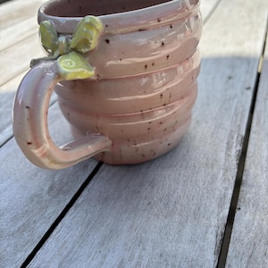 Könnte beinhalten: Rosa Keramikbecher mit geripptem Design und dekorativem Griff. Der Griff hat einen gelben floralen Akzent. Der Becher ist mit dunklen Flecken gesprenkelt und steht auf einer Holzoberfläche.