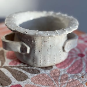 Könnte beinhalten: Eine handgefertigte, cremefarbene Keramikschale mit strukturierter Oberfläche und zwei kleinen Griffen. Der Rand hat einen einzigartigen, unebenen Rand. Die Schale ist mit kleinen braunen Punkten gesprenkelt und steht auf einer floral gemusterten Matte.