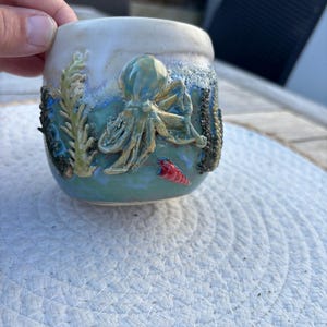Könnte beinhalten: Ein Keramikbecher mit einer erhabenen, dreidimensionalen Oktopus-Skulptur in Blau- und Grüntönen. Die Oberfläche zeigt strukturierte Algenmotive und eine kleine, leuchtend rote Muschel. Der obere Rand ist mit einer glatten, cremeweißen Glasur versehen.