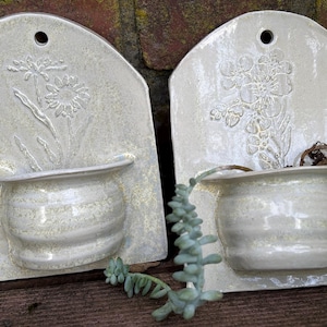 Könnte beinhalten: Zwei cremefarbene Wandtöpfe aus Keramik mit geprägten Blumendesigns. Jeder Pflanztopf hat einen kleinen, abgerundeten Topf und eine rechteckige Rückseite mit einem Loch zum Aufhängen. Einer zeigt eine Gänseblümchen, der andere einen Blumenstrauß.