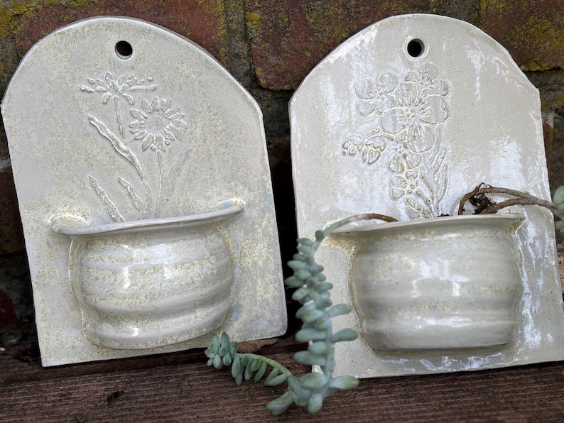 May include: Two off-white ceramic wall planters with embossed floral designs. Each planter has a small, rounded pot and a rectangular back with a hole for hanging. One planter features a daisy, the other a floral bouquet.