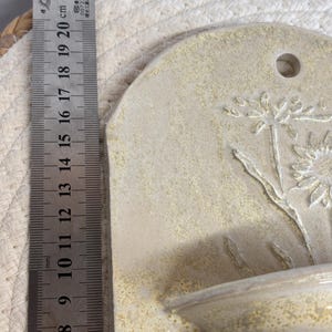 May include: A beige ceramic wall pocket with a floral design. The pocket has a small shelf and a hole for hanging. The ruler next to it measures up to 8 inches.
