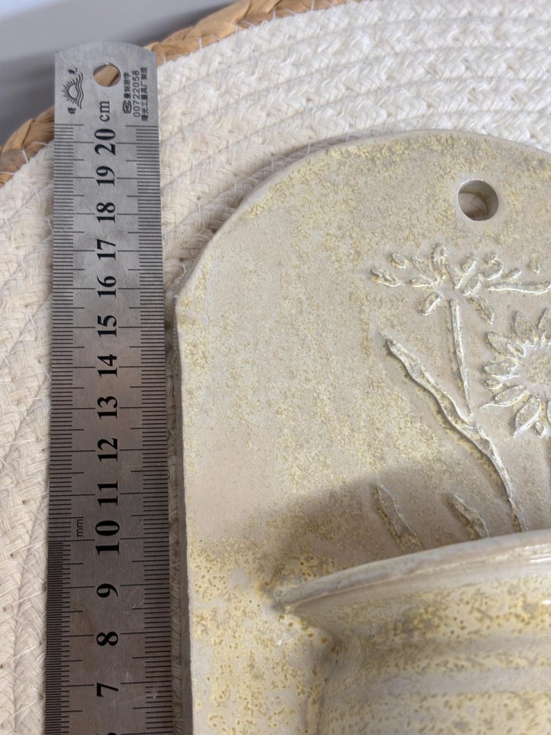 May include: A beige ceramic wall pocket with a floral design. The pocket has a small shelf and a hole for hanging. The ruler next to it measures up to 8 inches.