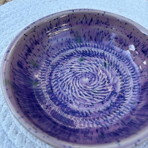 Könnte beinhalten: Eine runde Reibeschale aus Keramik mit einer glänzenden lavendelfarbenen Glasur und dunkelblauen Sprenkeln. Der innere Boden hat eine erhabene, spiralförmige Textur zum Reiben von Lebensmitteln, umgeben von einem glatten, breiten Rand.
