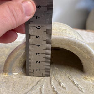 May include: A close-up of a beige ceramic lid with a handle. The handle's height is approximately 1.5 inches. The lid has a textured surface with a floral design. A metal ruler is used to measure the height.