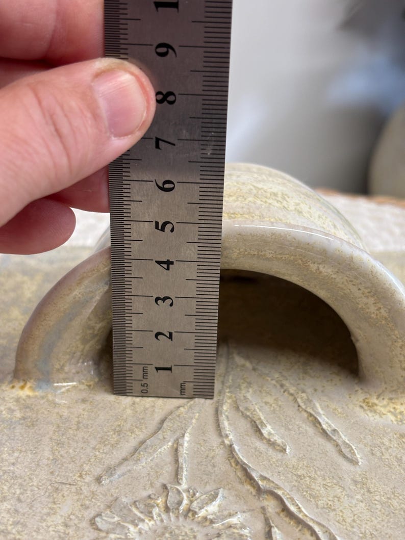 May include: A close-up of a beige ceramic lid with a handle. The handle's height is approximately 1.5 inches. The lid has a textured surface with a floral design. A metal ruler is used to measure the height.