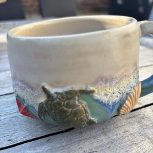 Könnte beinhalten: Ein Keramikbecher mit einem cremefarbenen oberen Teil und einem strukturierten unteren Bereich, der ein Relief einer grünen Meeresschildkröte, einer beigen Muschel und eines roten Seesterns zeigt. Der Bodenbereich zeigt Blau- und Grüntöne.