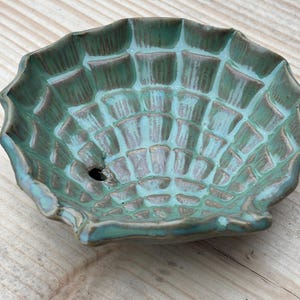 Könnte beinhalten: Eine keramische, muschelförmige Schale mit türkisfarbener und brauner Glasur. Die Schale hat ein strukturiertes, gitterartiges Muster und ein kleines Loch. Die Ränder sind gewellt, und die Schale steht auf einer Holzoberfläche.