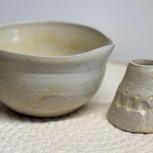 Könnte beinhalten: Eine Keramikschüssel mit Ausguss und eine kleine, strukturierte Vase. Die Schüssel und die Vase sind beide hellgrau mit gelben Sprenkeln. Die Vase hat eine strukturierte Oberfläche mit kreisförmigen Vertiefungen.