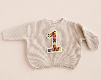 Puede incluir: Un suéter de punto beige con textura acanalada. El suéter presenta un gran número "1" bordado y colorido en la parte delantera, con un diseño floral en tonos amarillos, naranjas, morados y verdes. Tiene mangas largas y cuello redondo.