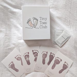 Puede incluir: Una caja rectangular blanca con la etiqueta Tiny Toes Club y un gráfico de huellas de pies, dos paquetes blancos pequeños y cuatro tarjetas blancas con huellas de bebé en rosa oscuro y el texto Hello Little, ONE, TWO, THREE.