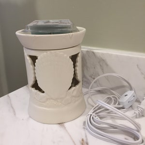 Puede incluir: Calentador de cera blanco de cerámica con un diseño decorativo de hojas y flores. El calentador tiene una parte superior redonda y un cable con un interruptor.