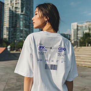 Könnte beinhalten: Weißes T-Shirt mit blauem Text und Grafiken. Das Design enthält die Texte "JFK NEW YORK" und "CDG PARIS" mit Flugdetails. Das T-Shirt hat eine lockere Passform.
