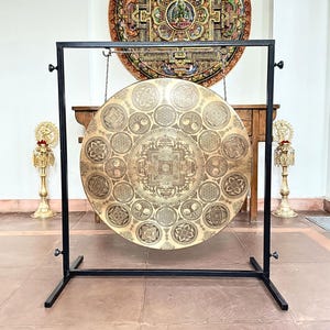 May include: A large circular brass gong suspended from a black metal frame. The gong surface features intricate etched geometric patterns, mandalas, and floral motifs arranged in concentric circles around a central square design element.