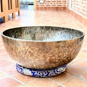 May include: A large, round, hammered bronze singing bowl with a dark, mottled patina and a circular etched symbol on the front. The bowl rests on a circular, blue and gold patterned fabric cushion with a floral and cloud-like design motif.