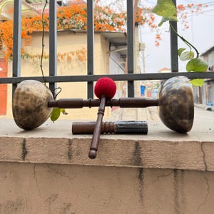May include: Two bronze-coloured singing bowls with wooden handles and a red mallet. The bowls are resting on a concrete ledge, with a second mallet and a small wooden cylinder also present. The background includes a metal fence and foliage.