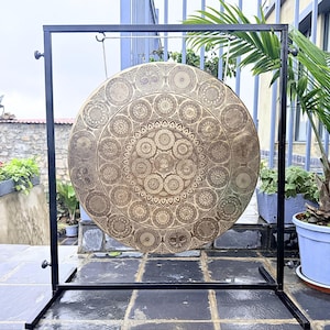 May include: A large, circular brass gong suspended from a black metal frame. The gong surface is intricately engraved with concentric rings of detailed geometric and floral patterns, featuring a central cluster of circular motifs and outer borders.
