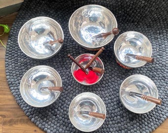 May include: A collection of silver-colored singing bowls with wooden strikers. The bowls are arranged on a dark gray, textured surface. One bowl contains red fabric. The wooden strikers have a dark brown finish.
