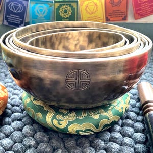 Puede incluir: Un juego de cuatro cuencos cantores de bronce de diferentes tamaños, anidados entre sí. Cada cuenco tiene un emblema decorativo. Los cuencos descansan sobre un cojín verde y dorado. Un mazo de madera es visible.