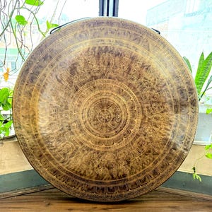 May include: A large, circular bronze gong with detailed etched patterns. The gong has concentric circles and ornate designs, possibly for meditation or sound healing. The dark bronze colour and carvings suggest a cultural or spiritual significance.