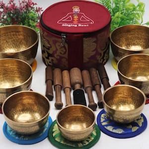 Puede incluir: Un juego de siete cuencos tibetanos de latón con baquetas de madera, dispuestos alrededor de un estuche de transporte rojo. El estuche tiene una cremallera y un diseño con una figura y las palabras "Singing Bowl". Los cuencos están sobre cojines redondos coloridos.