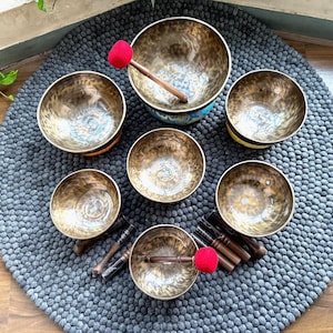 Puede incluir: Un conjunto de siete cuencos cantores de color bronce de varios tamaños, dispuestos sobre una alfombra circular gris texturizada. Cada cuenco tiene un patrón único. Se incluyen mazos de madera con superficies de golpeo de fieltro rojo.