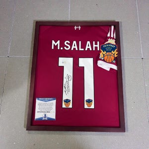 May include: Framed burgundy jersey with the name "M. SALAH" and the number "11" in white. The jersey is signed and includes a "TRIKINGS CUP" patch and a Beckett Certificate of Authenticity. The frame is a dark brown color.