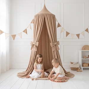 May include: A beige canopy bed with large bows and a matching garland of triangular flags. Two children are playing with wooden toys inside the canopy. The room has white walls and a white wooden floor.