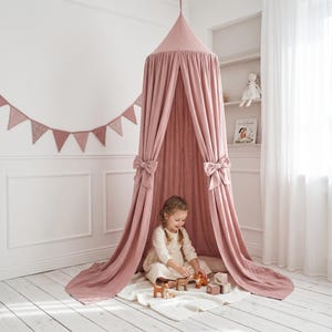 May include: A dusty rose canopy bed with draped fabric and bow accents. A young child is playing with wooden toys inside the canopy. A matching garland of pennants hangs on the wall.
