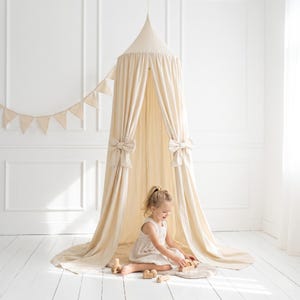 May include: A cream-colored canopy bed with a conical top and draped fabric, adorned with large bow accents. A small garland of triangular flags hangs nearby. A young child is playing with wooden toys inside the canopy.