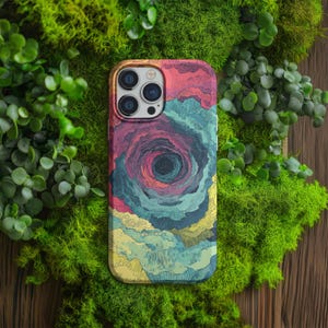 May include: A mobile phone case with a vibrant, abstract spiral design in red, blue, yellow, and purple hues. The case has a glossy finish and a camera aperture. The case is surrounded by green moss and foliage.