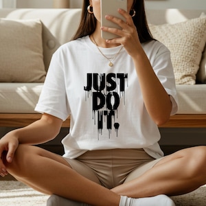 Puede incluir: Camiseta blanca con la frase "JUST DO IT" en letras negras goteantes. La camiseta de manga corta se combina con pantalones cortos beige. La persona est&aacute; sentada en una alfombra de color claro. El estilo es casual.