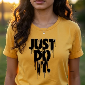 Puede incluir: Camiseta amarilla mostaza con mangas enrolladas. El texto negro "JUST DO IT." est&aacute; impreso en la parte delantera con un efecto de goteo. La camiseta est&aacute; hecha de un material suave.